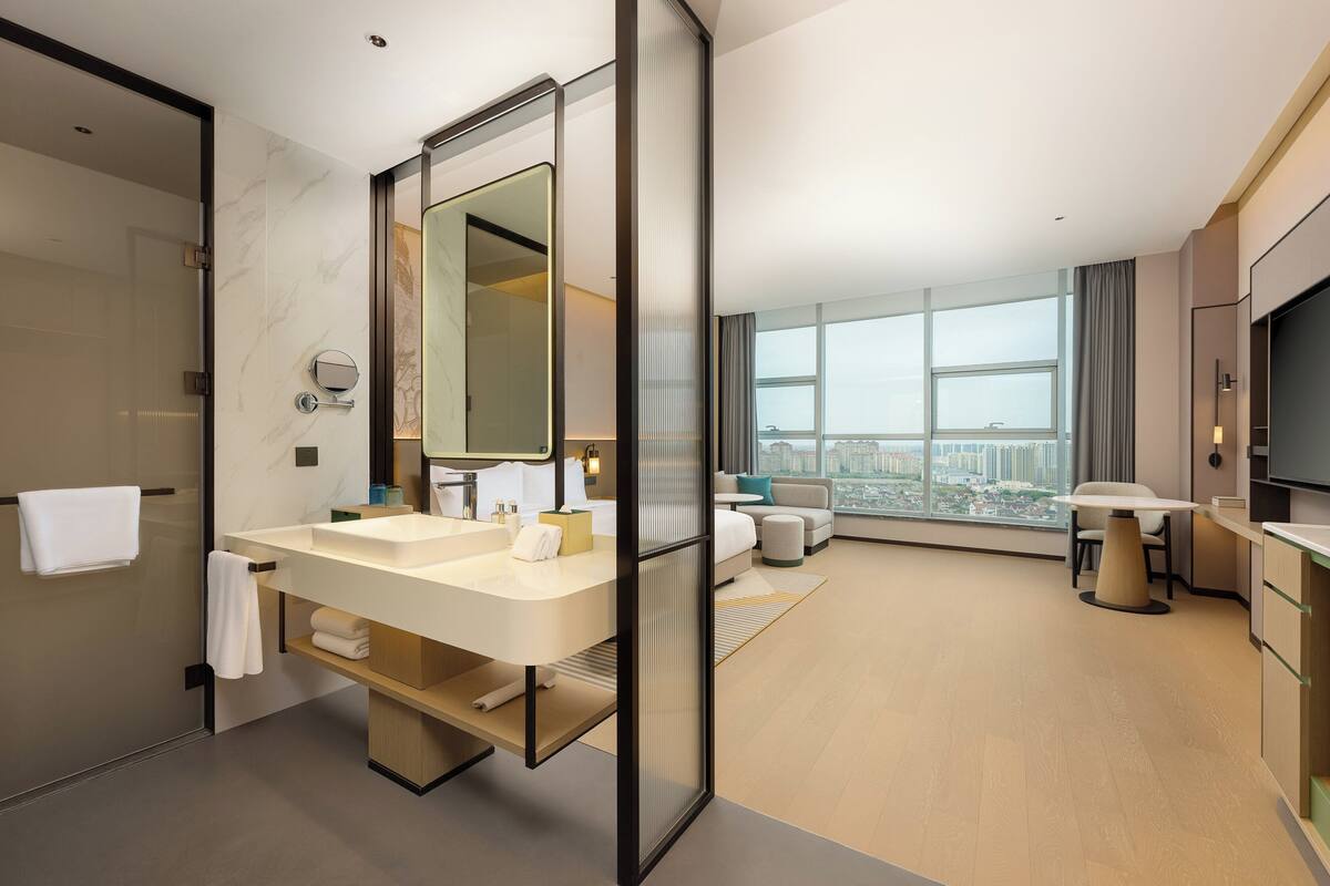 Un baño de hotel moderno con lavamanos dobles, un espejo grande y vistas a la ciudad a través de ventanales amplios.