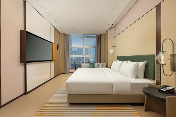 Ein Hotelzimmer mit einem großen Bett, einem Flachbildfernseher, einem Schreibtisch mit Telefon und Blick auf die Stadt durch das Fenster.