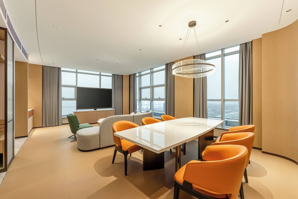 Una habitación de hotel moderna con un ventanal grande, un televisor de pantalla plana, una mesa de comedor con sillas naranjas y un sofá.