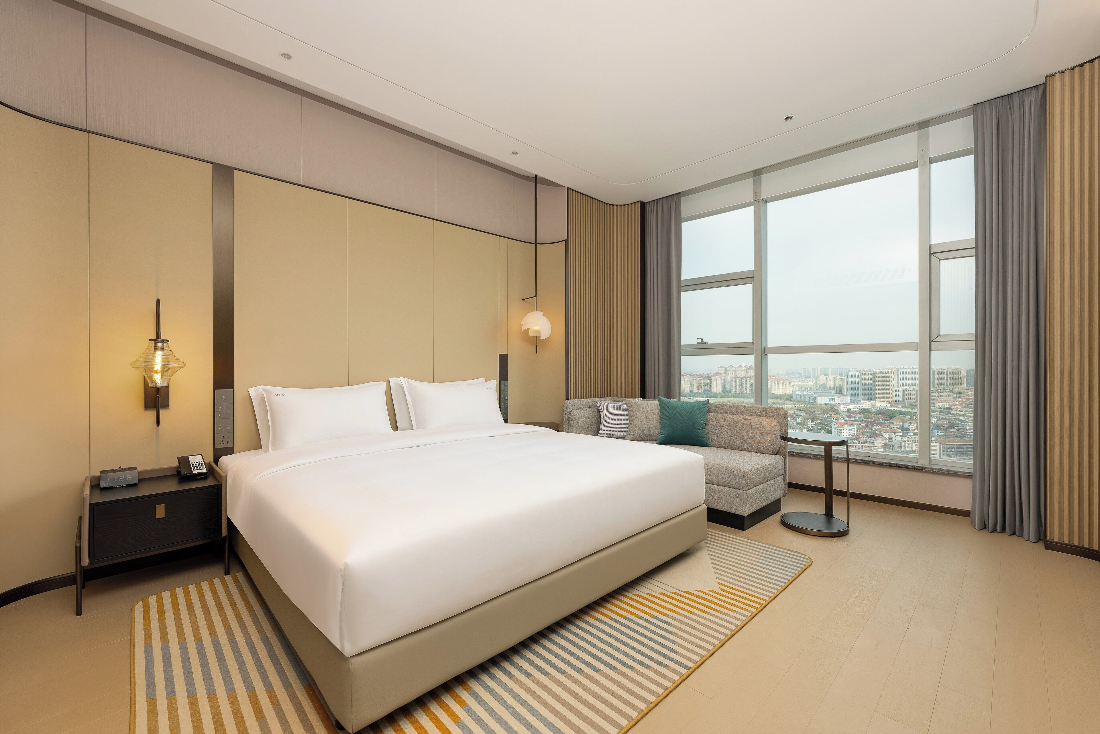 A modern hotel room with a large bed, a sofa, a nightstand, a lamp, and a view of the city through large windows.