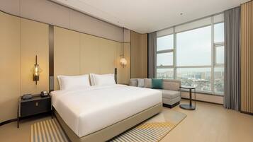 A modern hotel room with a large bed, a sofa, a nightstand, a lamp, and a view of the city through large windows.