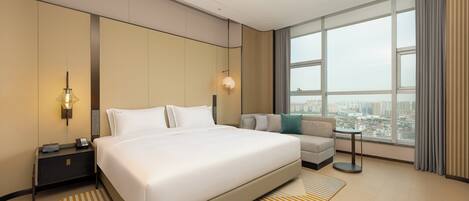 Habitación de hotel moderna con una cama grande, un sofá, una mesita de noche, una lámpara y vistas de la ciudad a través de amplios ventanales.