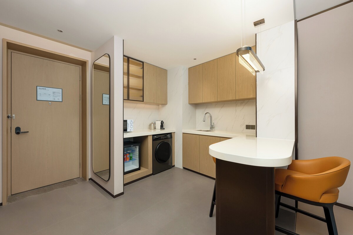 Habitación de hotel compacta con kitchenette, lavadora, lavamanos, espejo y una sola silla naranja.