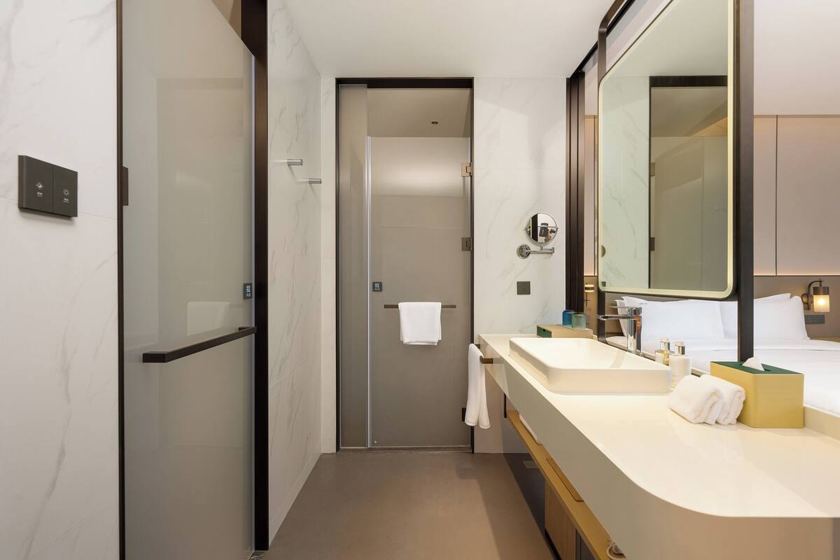 Un baño de hotel moderno con cabina de ducha de vidrio, tocador con lavabo y espejo, y una cama con ropa blanca.