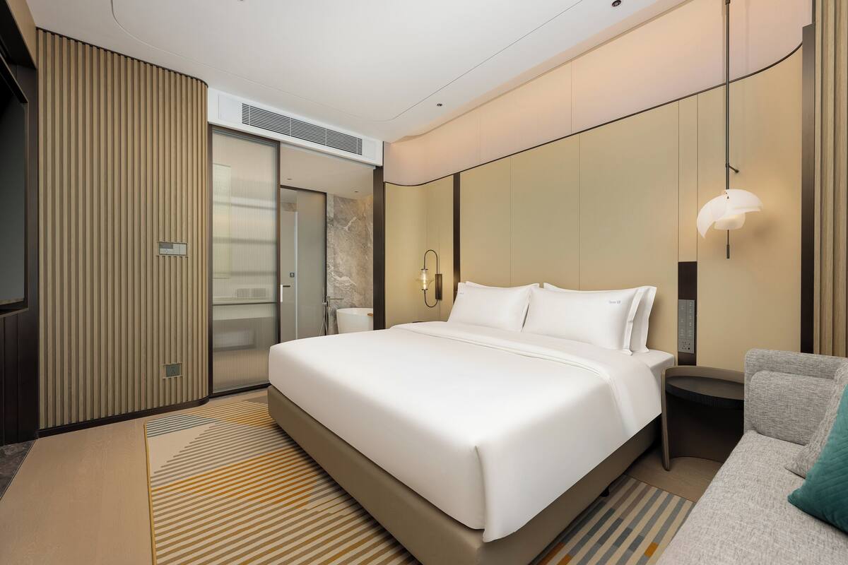 Una habitación de hotel moderna con una cama grande, una mesita de noche, un sofá, un televisor montado en la pared y un baño con ducha e inodoro.