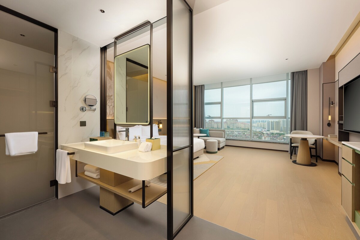 Un baño de hotel moderno con lavamanos dobles, un espejo grande y vistas a la ciudad a través de ventanales amplios.