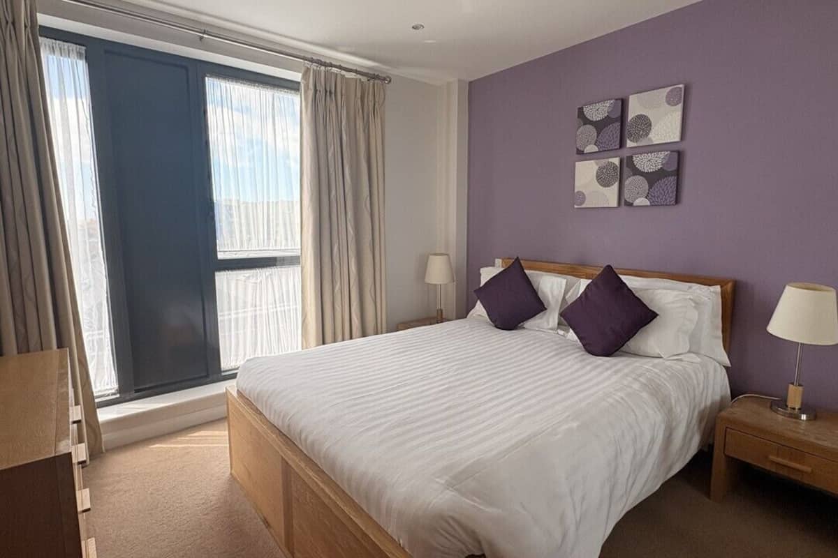 A hotel room with a bed, a nightstand, a window with curtains, and wall art.