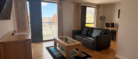 A modern living room with a sofa, a glass-top coffee table, and a desk with a chair. Large windows offer a view of the cityscape.