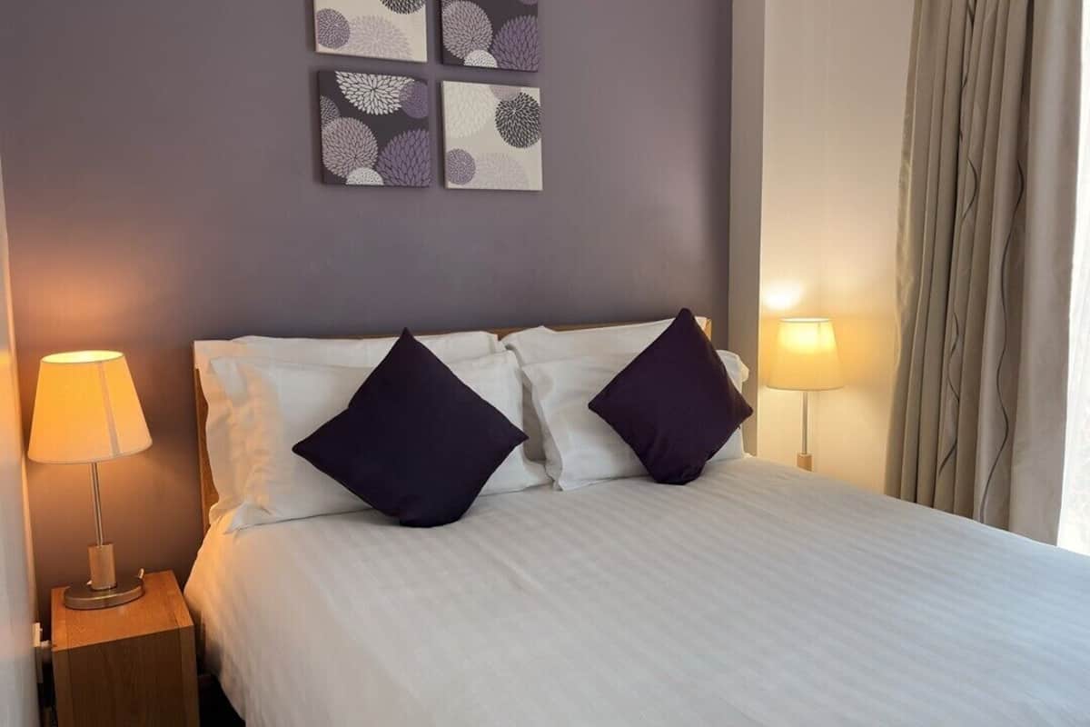 A neatly made bed with white linens and two dark pillows, flanked by two bedside lamps on wooden nightstands.
