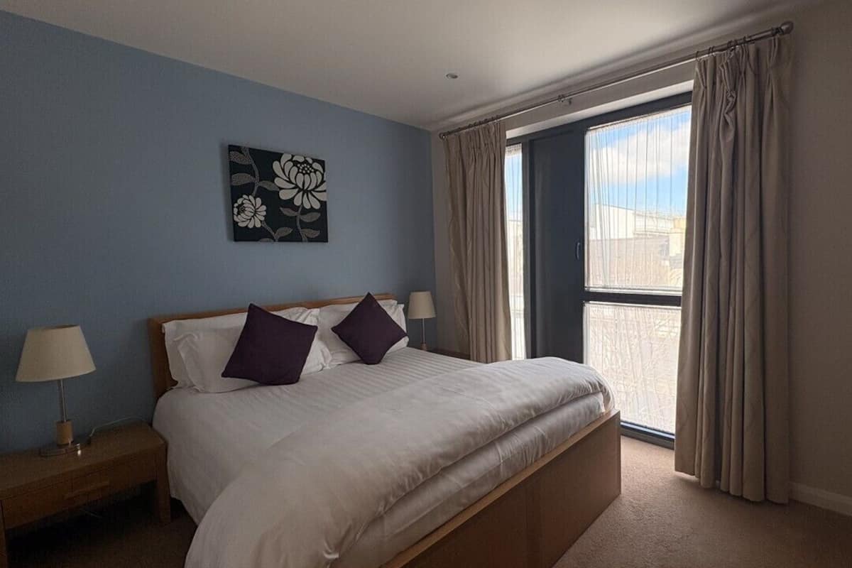 A bedroom with a bed, two pillows, a nightstand, a lamp, a window with curtains, and a wall art piece.