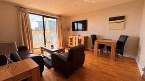 A modern living room with a sofa, a dining table with chairs, a TV mounted on the wall, and a balcony with curtains. - Liberty Wharf Apartments (St. Helier)
