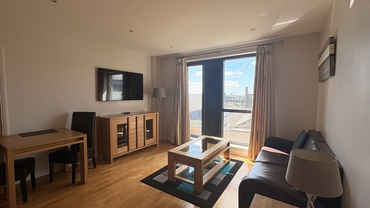 A modern living room with a wooden table, a sofa, a TV mounted on the wall, and a sliding glass door leading to a balcony.