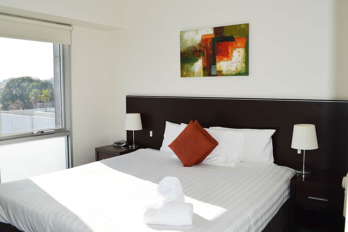 Une chambre d’hôtel avec un grand lit, du linge de lit blanc, un oreiller rouge, deux lampes de chevet, un tableau au mur et une fenêtre donnant sur des arbres.