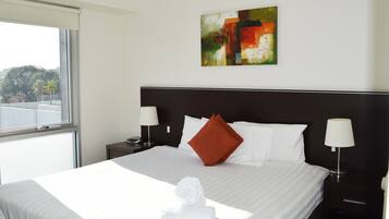 Una habitación de hotel con una cama grande, ropa de cama blanca, una almohada roja, dos lámparas de noche, un cuadro en la pared y una ventana con vistas a árboles.
