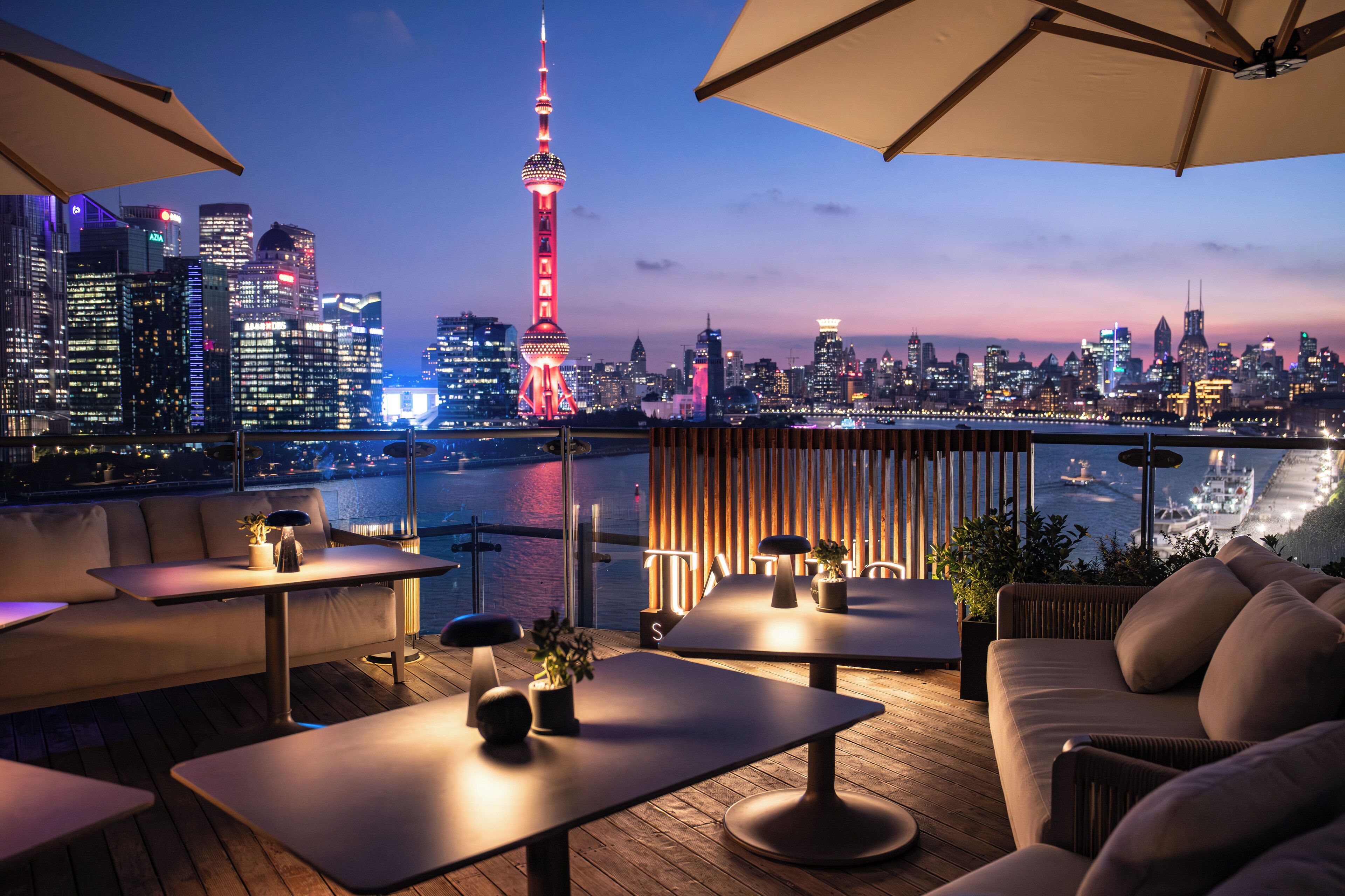 Une terrasse sur le toit, dotée de tables et de sièges, offrant une vue sur les toits de la ville et sur une étendue d’eau au crépuscule.