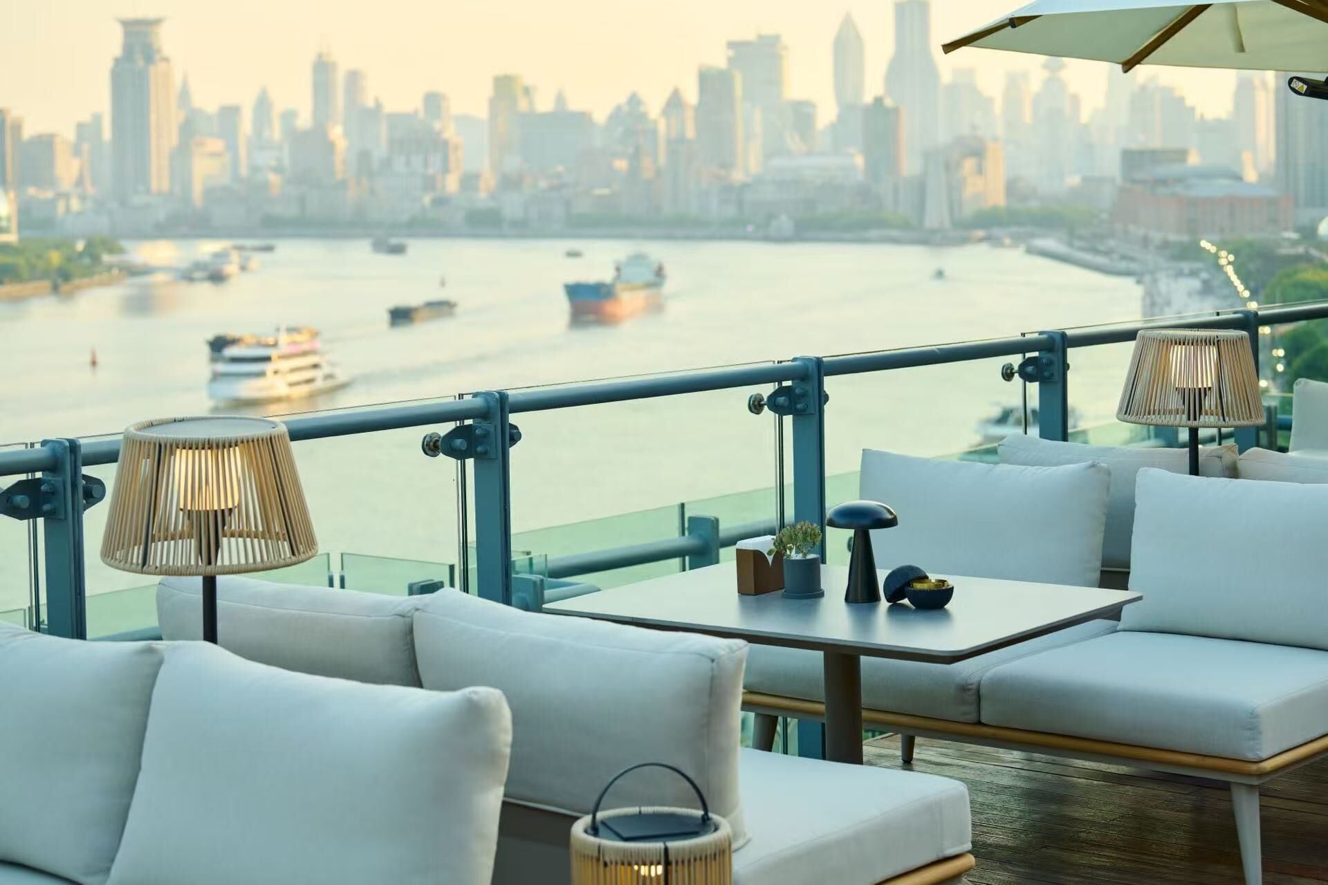Un área de estar moderna al aire libre con sofás blancos y mesas, con vistas a un río donde navegan barcos y un horizonte urbano al fondo.