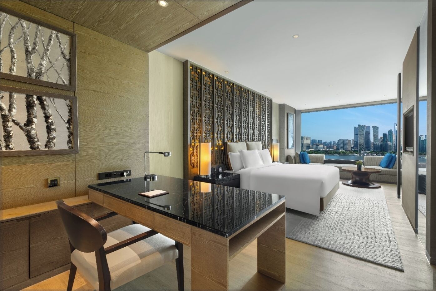 A modern hotel room with a city view, a large bed, a desk, and a seating area.
