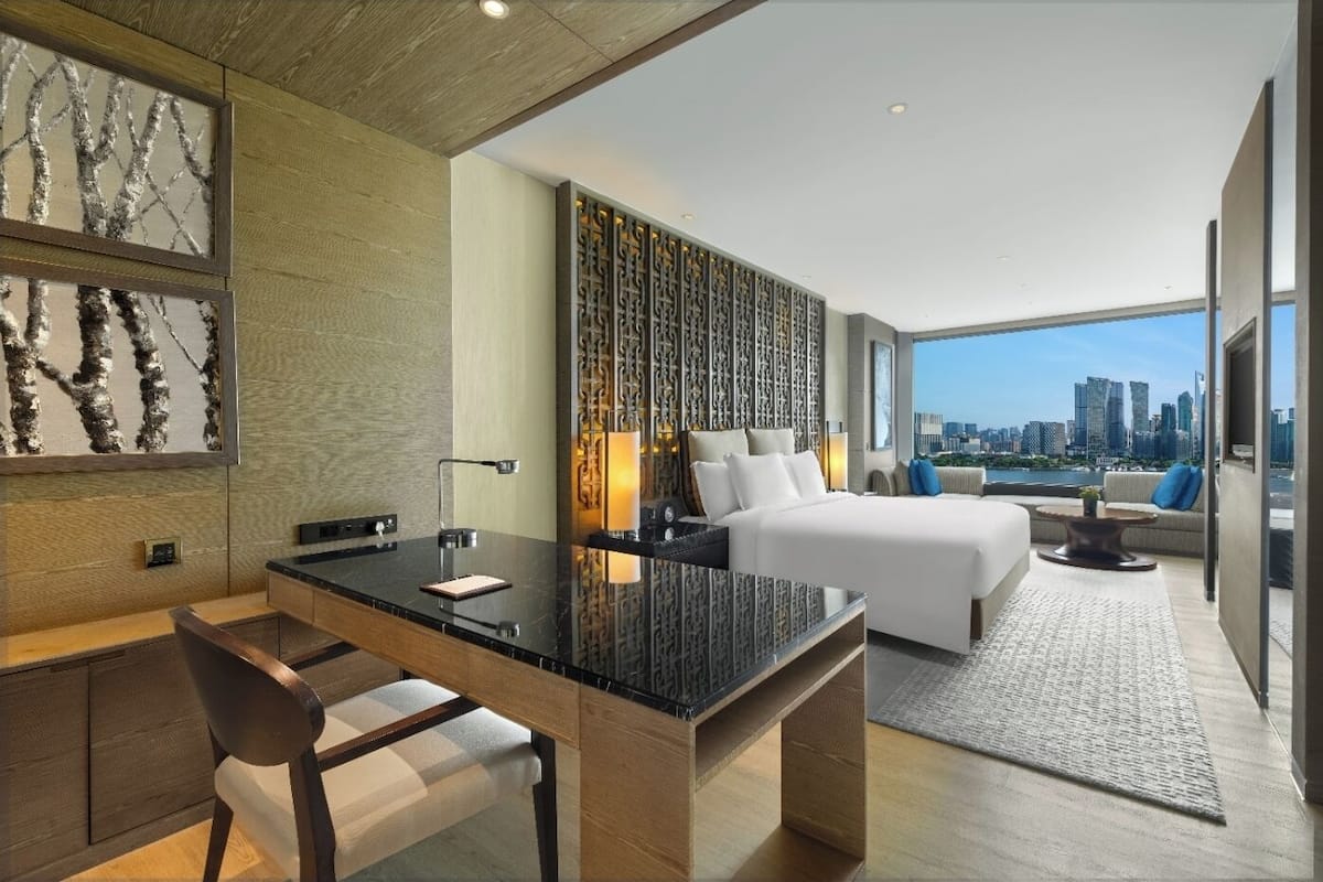 A modern hotel room with a city view, a large bed, a desk, and a seating area.