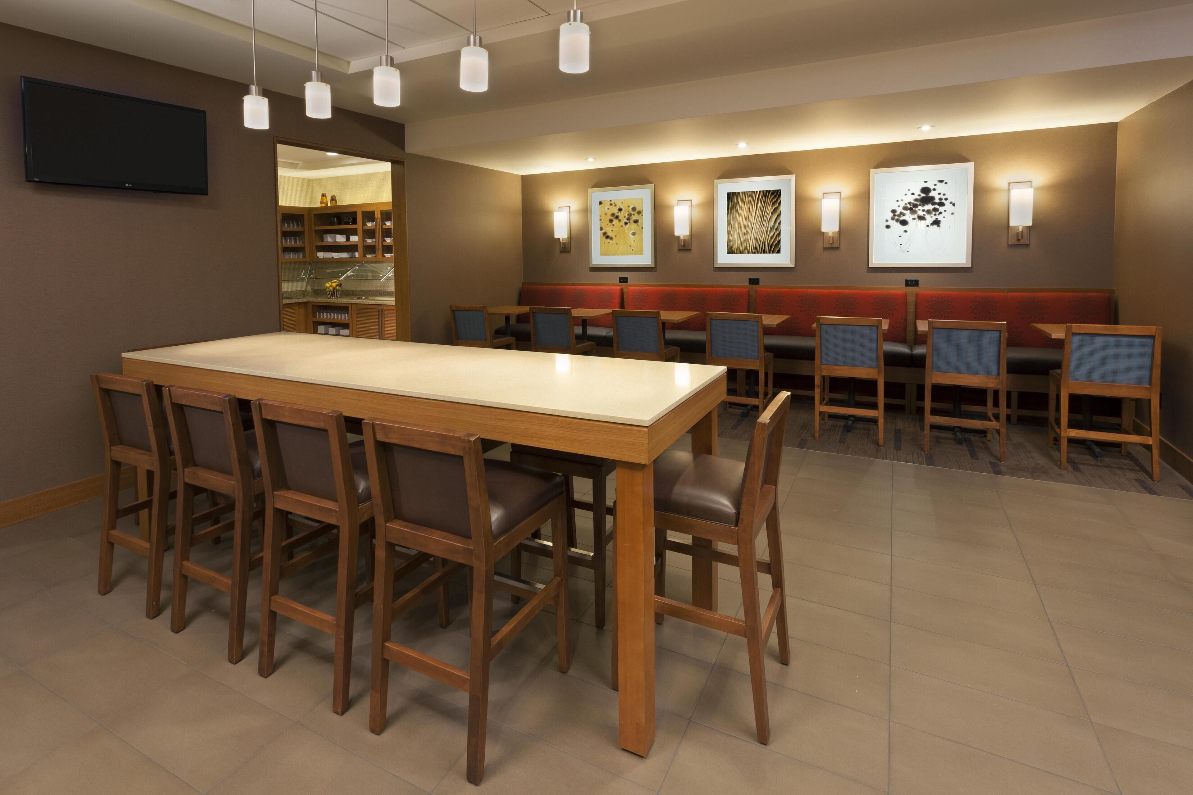 A hotel dining area with a large wooden table, multiple chairs, and wall-mounted artwork.