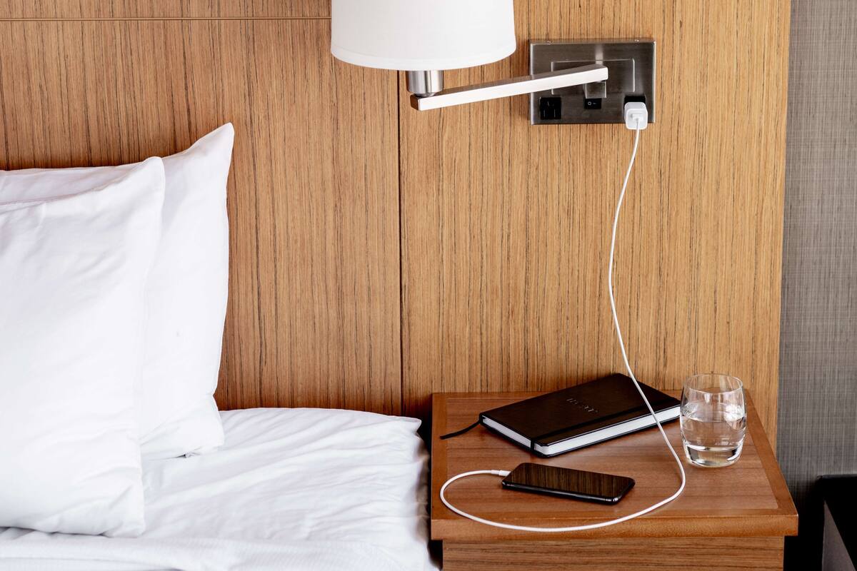 A neatly made bed with white linens, a bedside table with a lamp, a notebook, a smartphone, and a glass of water.