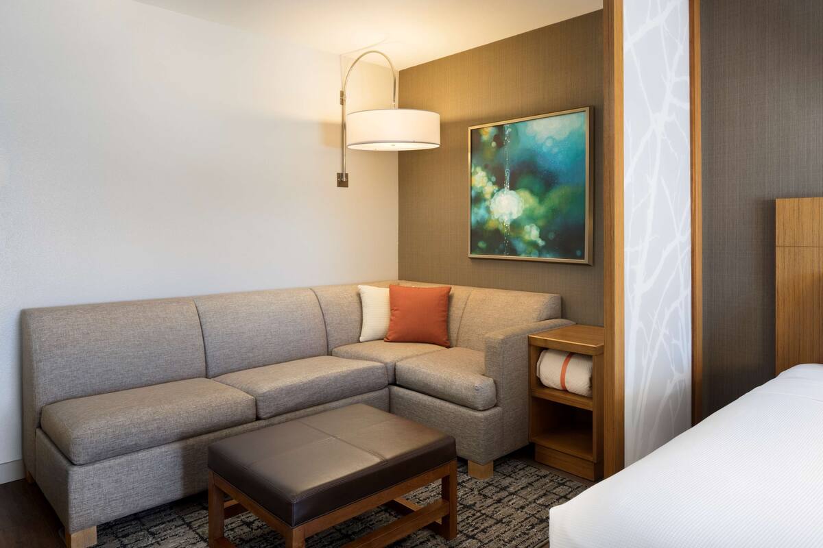 A modern hotel room with a sectional sofa, a wooden coffee table, a bed, a painting on the wall, and a lamp.
