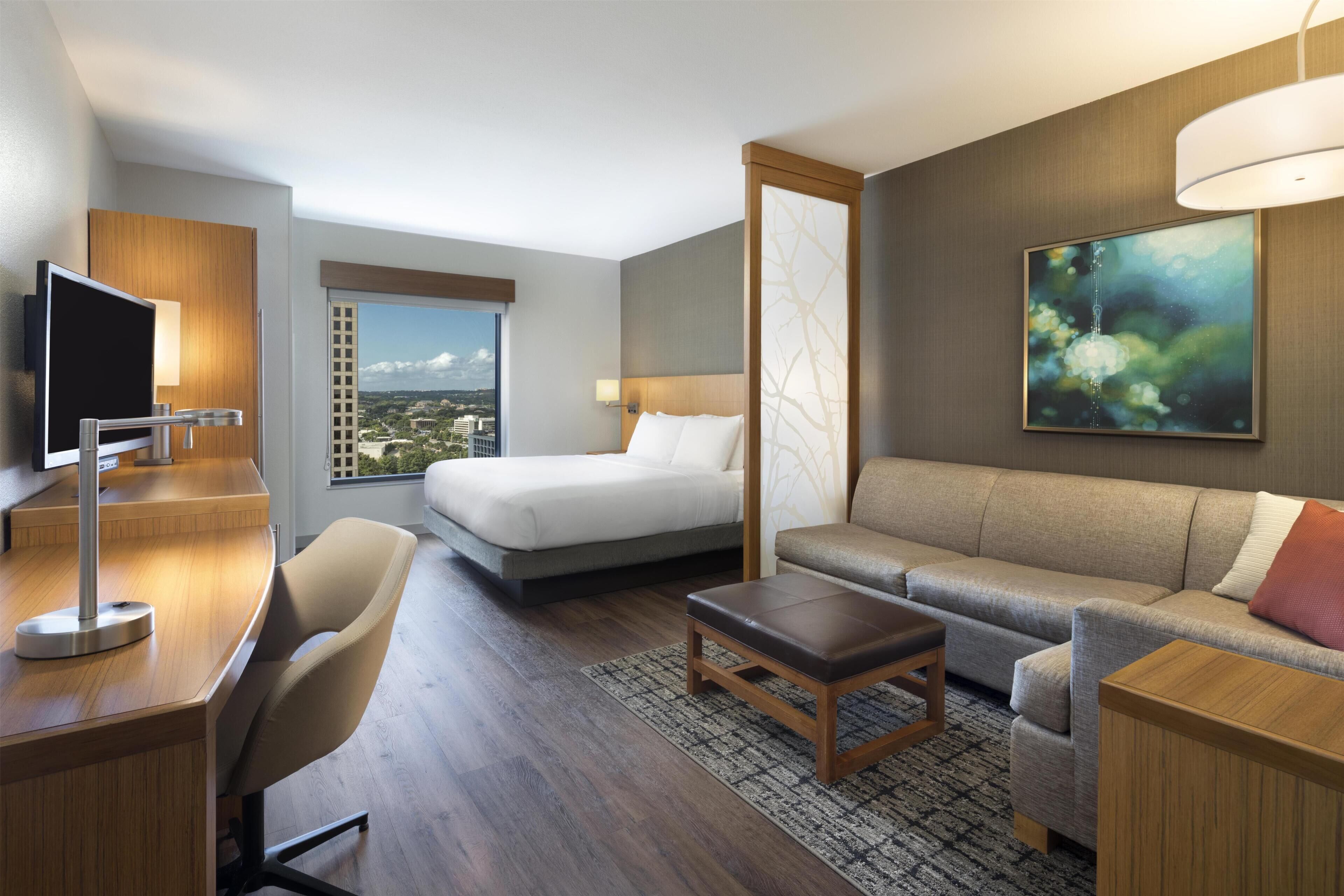 A hotel room with a bed, sofa, desk, TV, and a large window with a city view.