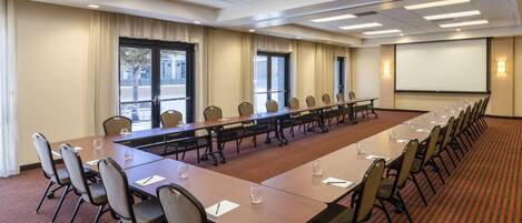 Un salón de conferencias con una larga mesa, sillas, una pantalla de proyector y ventanas.
