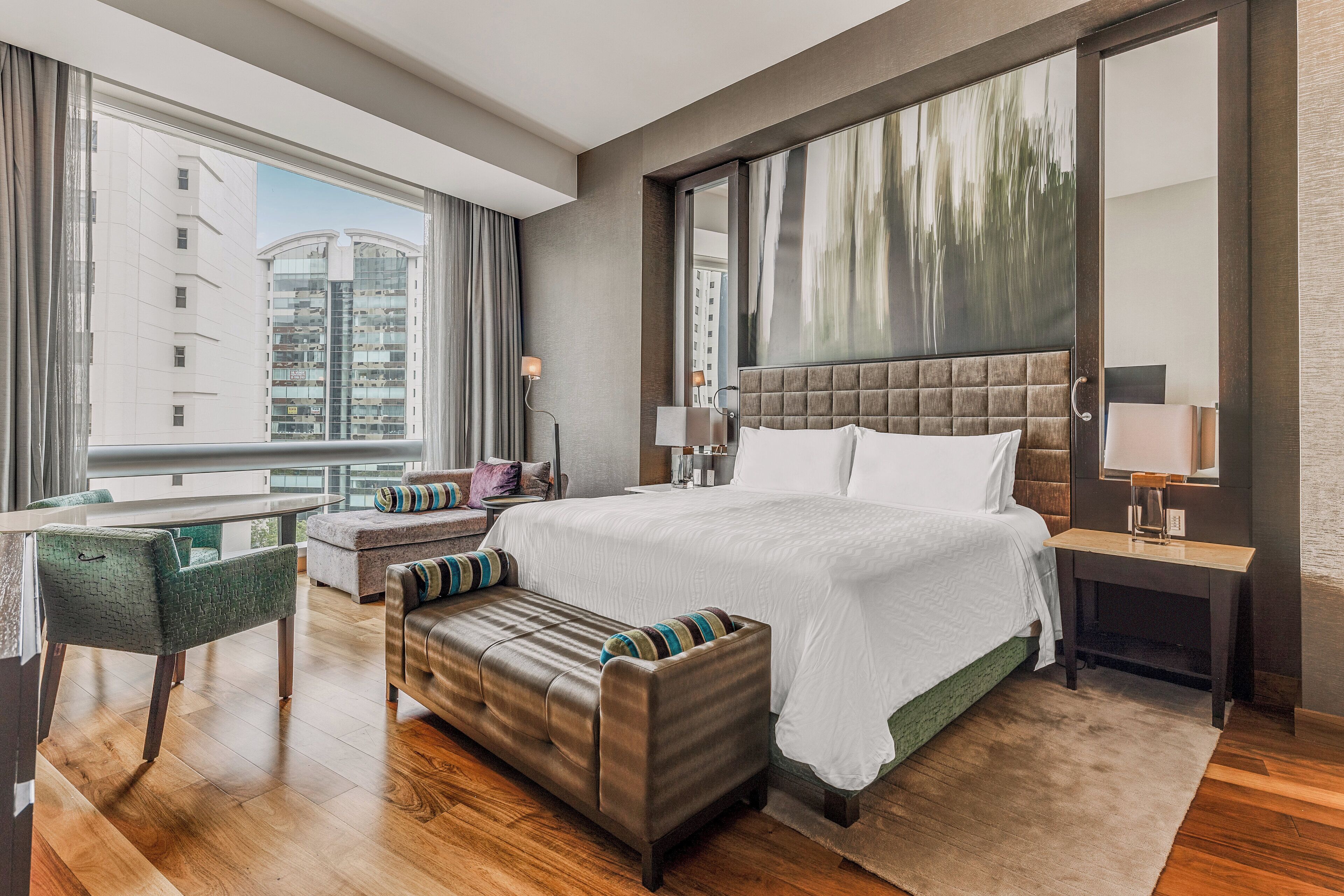 A modern hotel room with a large bed, a seating area with a bench, a desk, and a large window offering a city view.