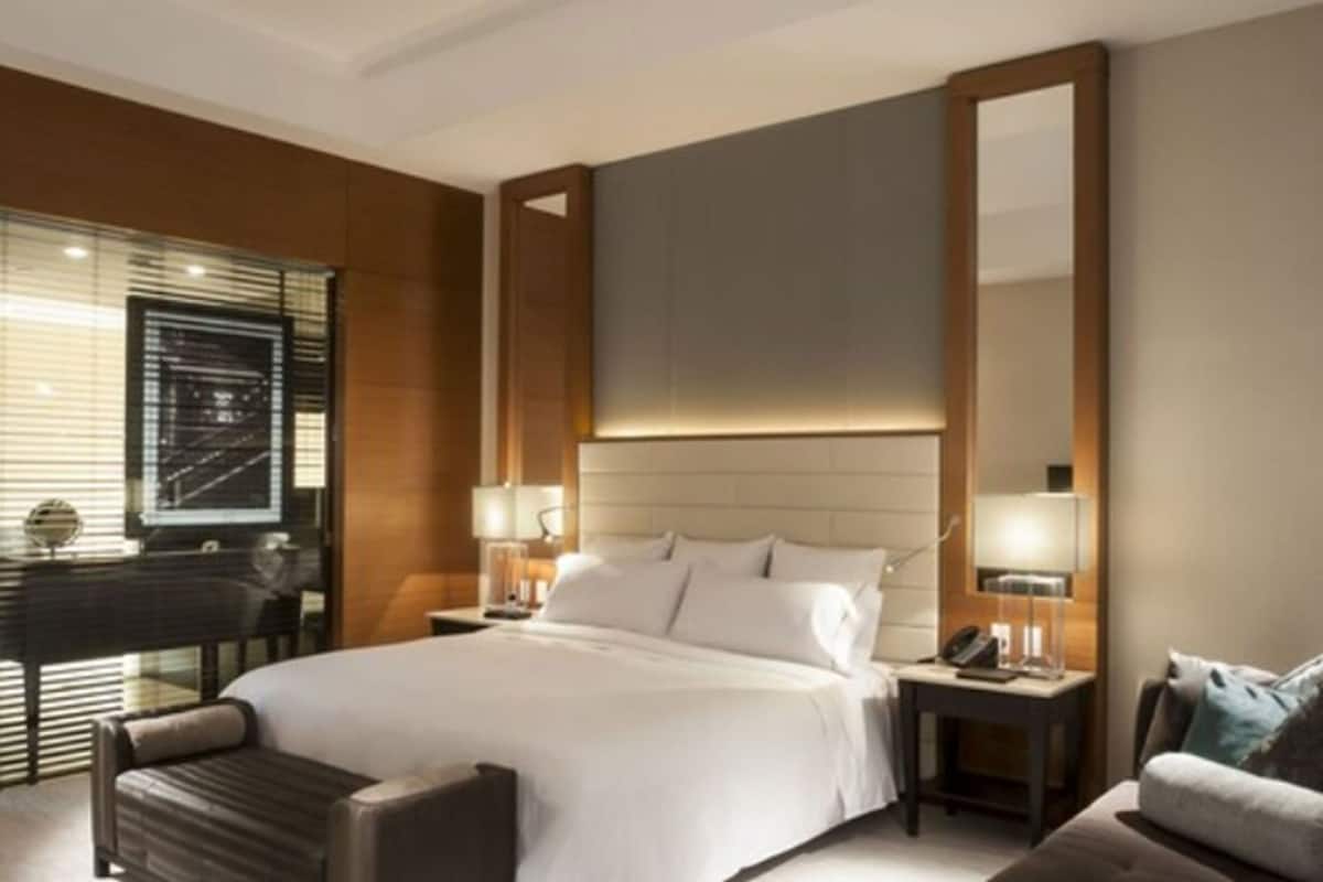 Una habitación de hotel moderna con una cama grande, dos mesitas de noche con lámparas, un área de descanso con sofá y un espejo amplio.