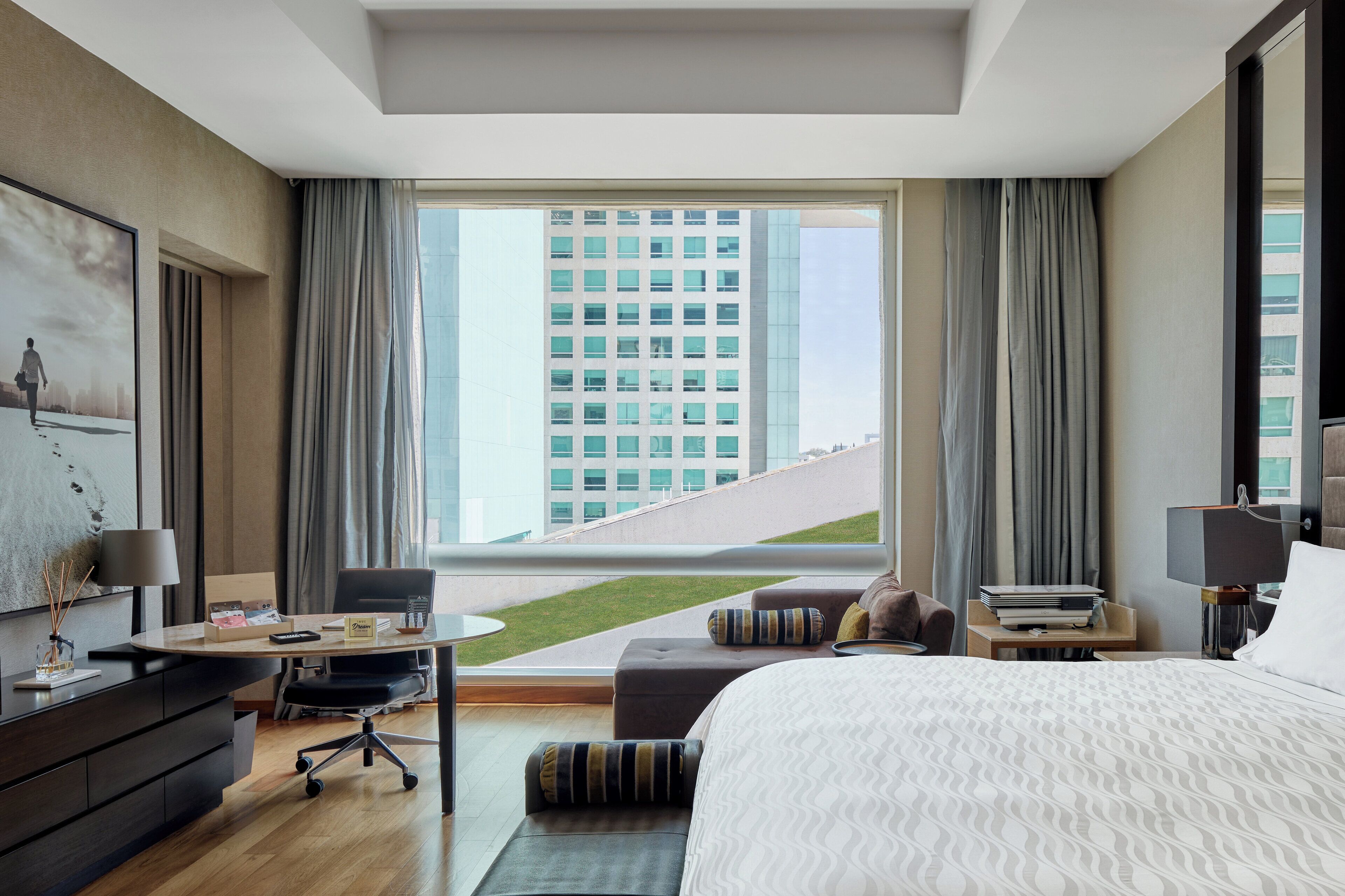 Una habitación de hotel moderna con una cama grande, un escritorio, una silla y vistas a un paisaje urbano a través de amplios ventanales.