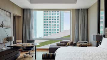 Una habitación de hotel moderna con una cama grande, un escritorio, una silla y vistas a un paisaje urbano a través de amplios ventanales.