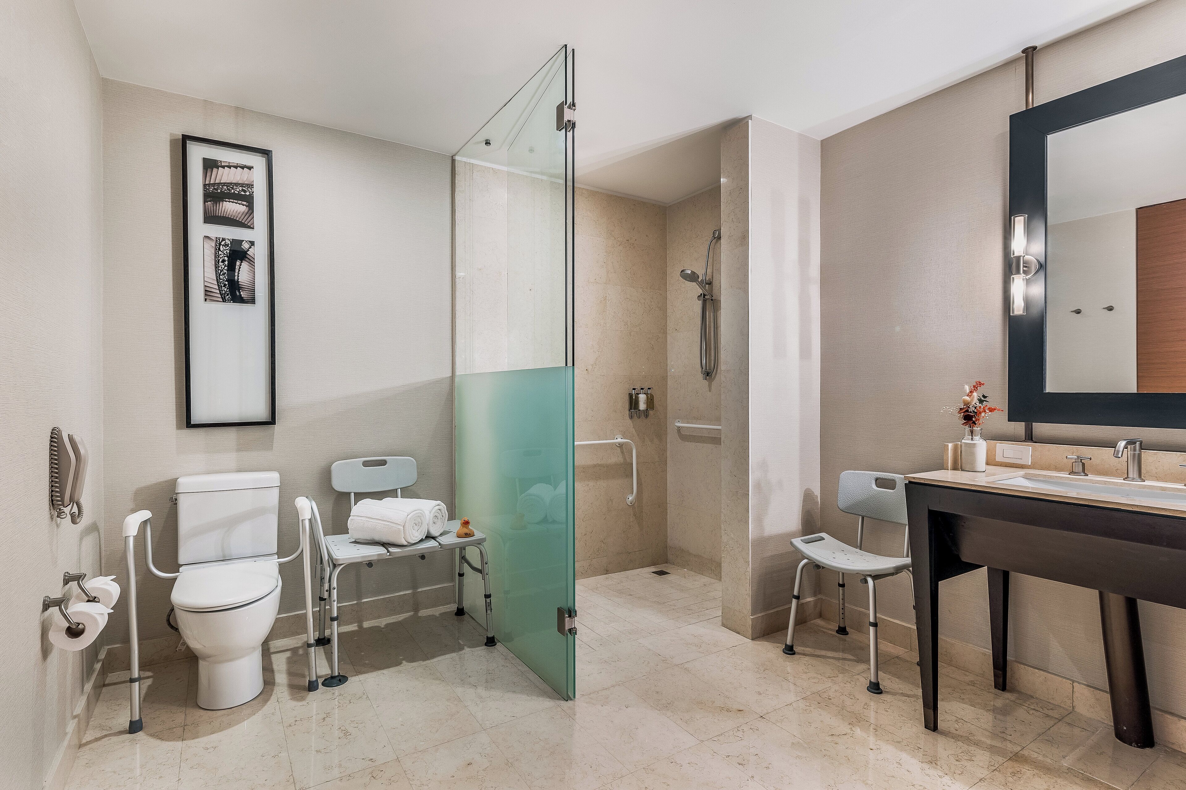 A modern bathroom with a glass shower enclosure, a white toilet, a sink with a mirror, and a wheelchair-accessible chair.