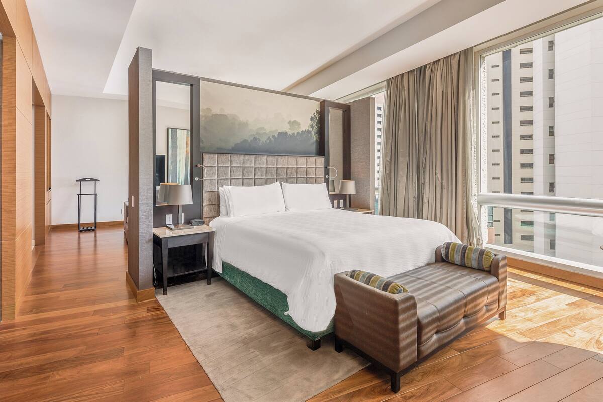 Ein modernes Hotelzimmer mit einem großen Bett, einer Sitzecke mit Sofa, einem Nachttisch, einer Lampe und einem großen Fenster mit Blick auf ein Gebäude.