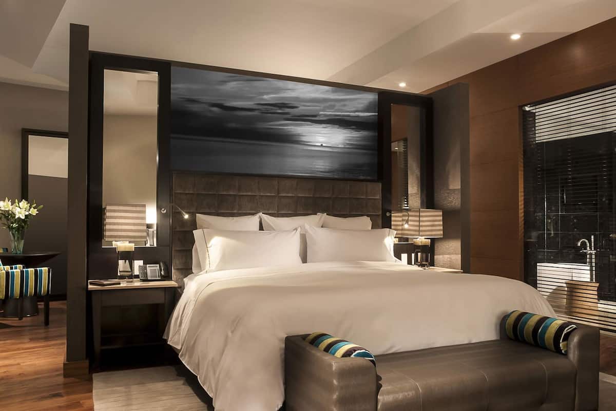 Un dormitorio moderno con una cama grande, una mesita de noche, un banco y un amplio ventanal con persianas.