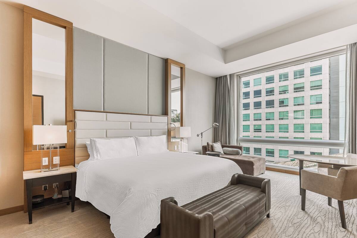 Ein Hotelzimmer mit einem großen Bett, einem Schreibtisch, einem Sessel und einem Spiegel, mit Blick auf ein Gebäude.