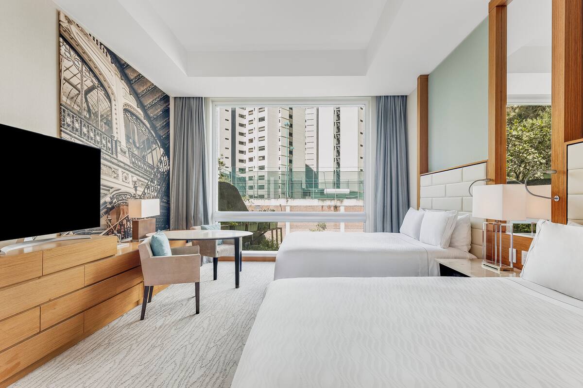 Ein Hotelzimmer mit einem großen Bett, einem Fernseher, einem Schreibtisch und einem Balkon mit Blick auf die Stadt.