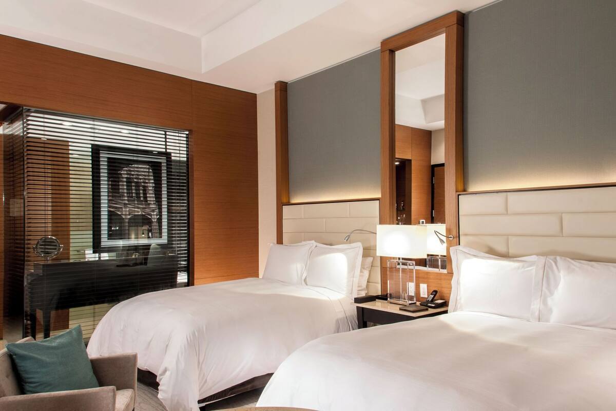 Habitación de hotel con dos camas, un escritorio, una silla, un espejo grande y una ventana con persianas.
