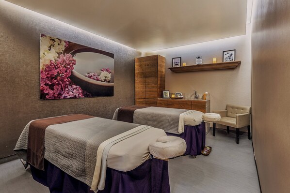 A spa room with two massage beds, a wooden cabinet, a chair, and a shelf with candles and pictures.