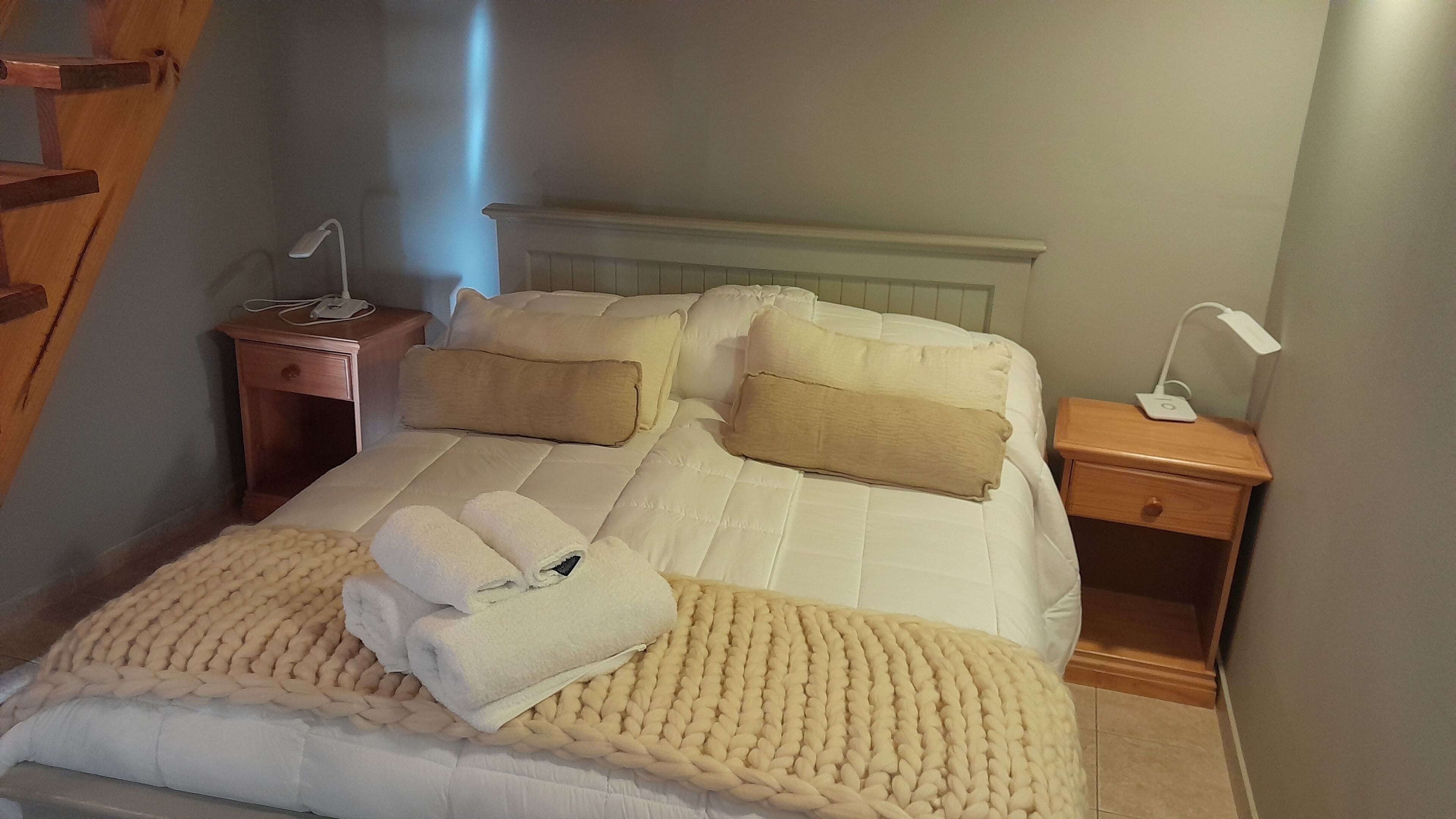 A neatly made bed with a white comforter, two beige pillows, and a beige knitted blanket. There are two bedside tables, each with a lamp and a phone.