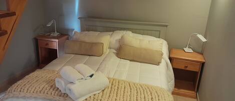 A neatly made bed with a white comforter, two beige pillows, and a beige knitted blanket. There are two bedside tables, each with a lamp and a phone.