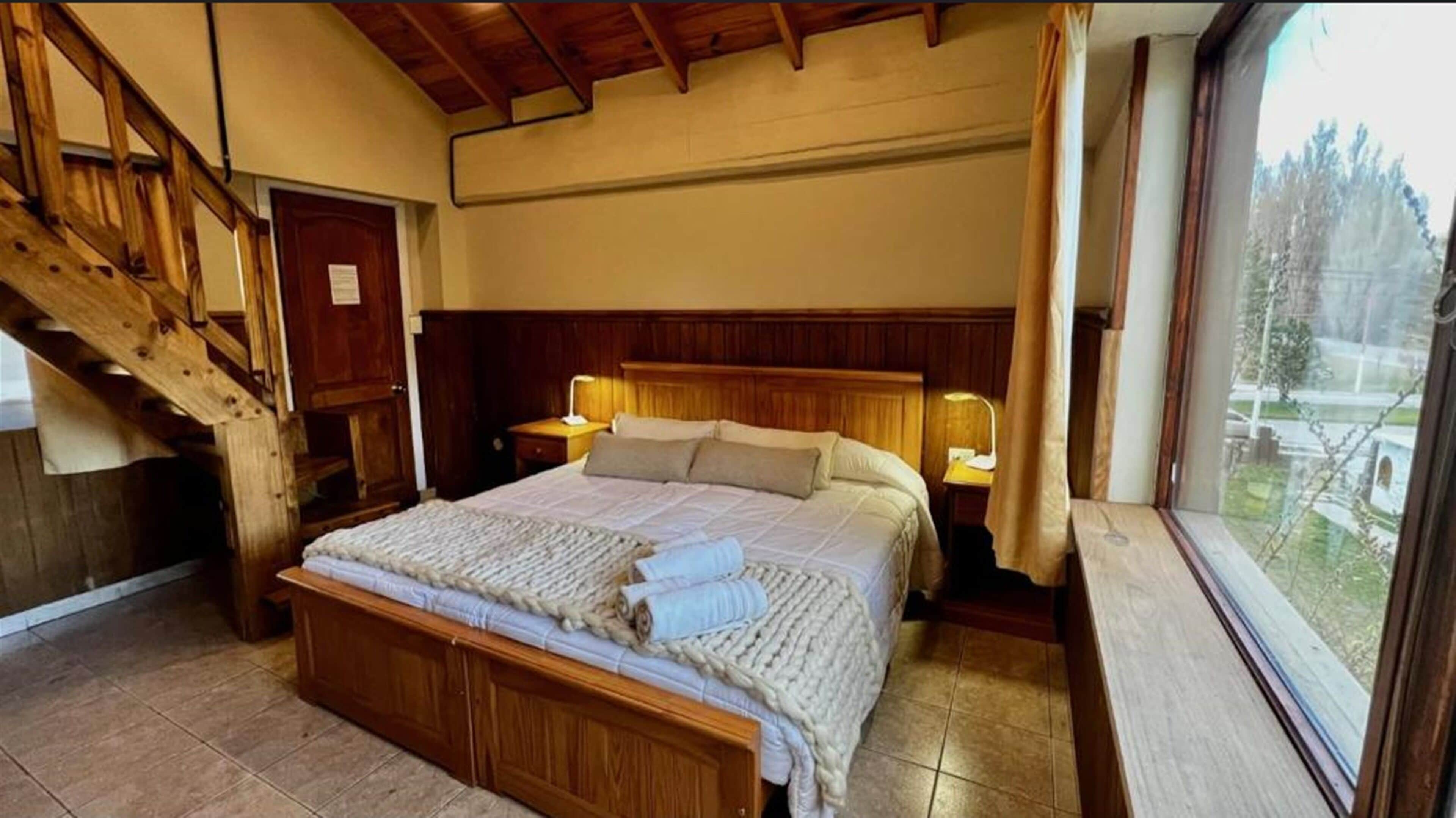 Ein Schlafzimmer mit einem hölzernen Bett, einem Nachttisch, einer Lampe, einem Fenster mit Aussicht und einer Treppe.