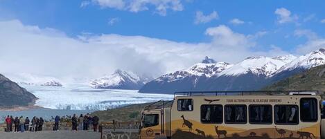 Un autobús turístico estacionado cerca de un glaciar, con montañas al fondo.