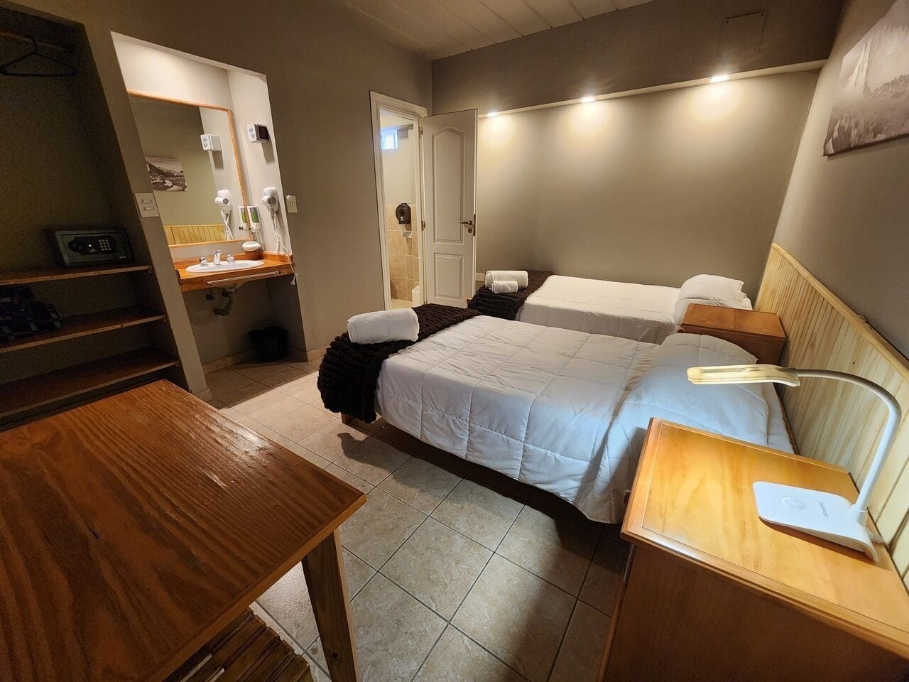 A compact hotel room with a bed, bedside table, and a small desk with a lamp.