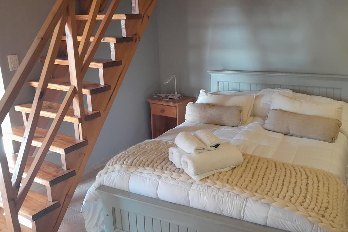 A bedroom with a wooden ladder, a bed with white bedding, a bedside table with a lamp, and a nightstand with a phone.