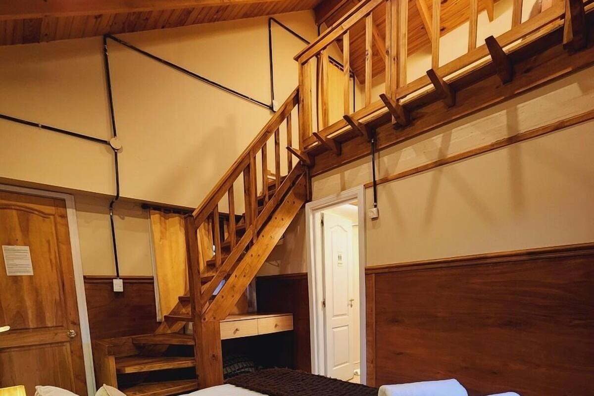 A wooden staircase leading to a loft area, a bed with white bedding and pillows, and a door with a light switch.