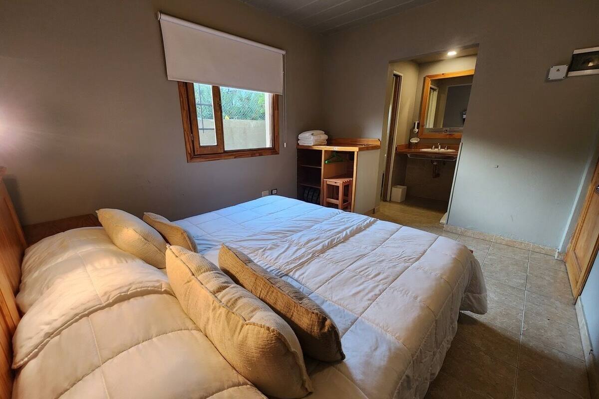 A bedroom with a large bed, a window with a blind, a desk with a chair, and a bathroom visible through an open door.
