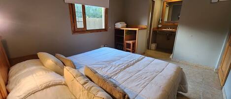 Un dormitorio con una cama grande, una ventana con persiana, un escritorio con silla y un baño visible a través de una puerta abierta.
