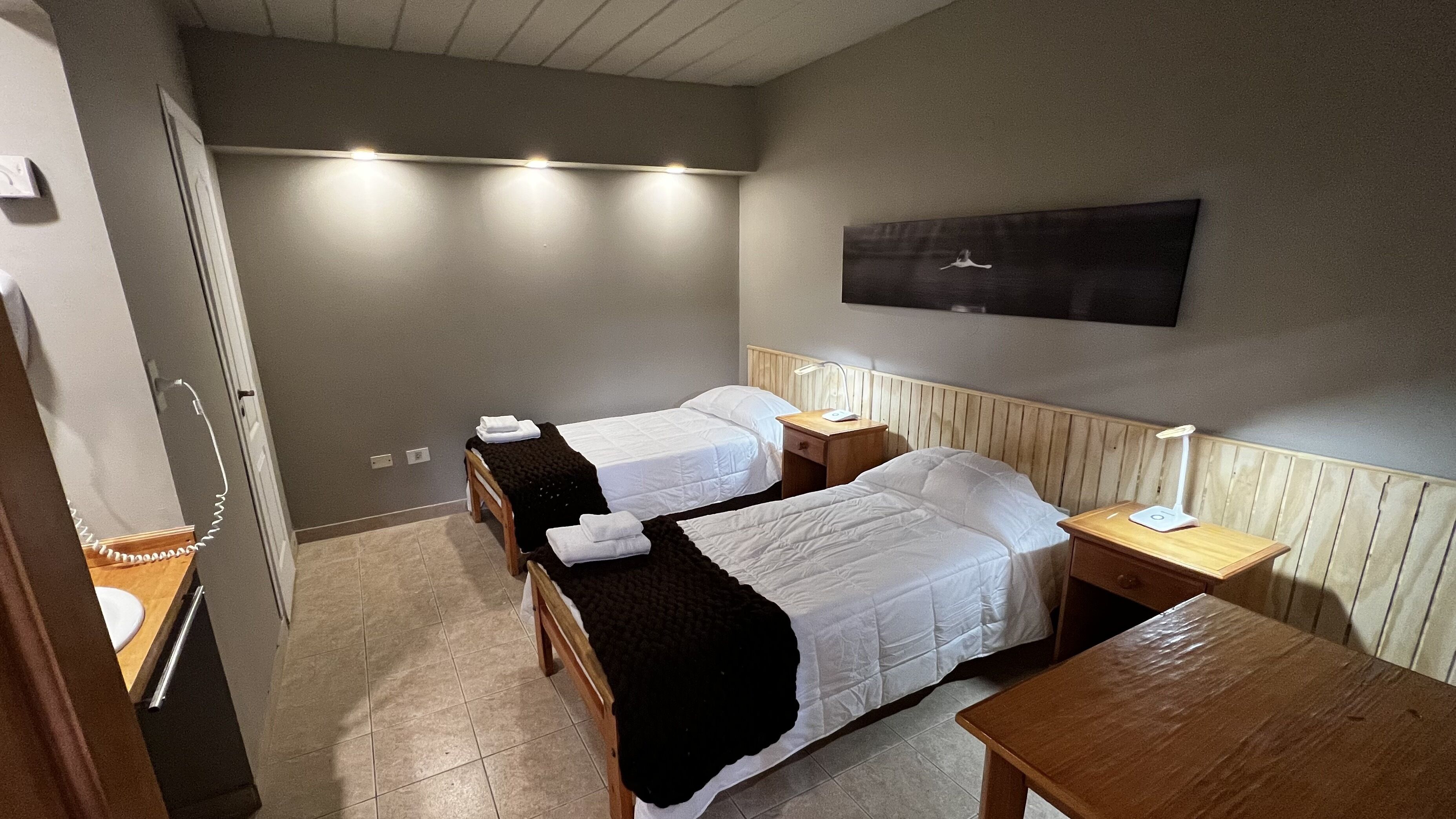 A hotel room with two single beds, a wooden nightstand with a lamp, a wooden table, and a wall-mounted TV.