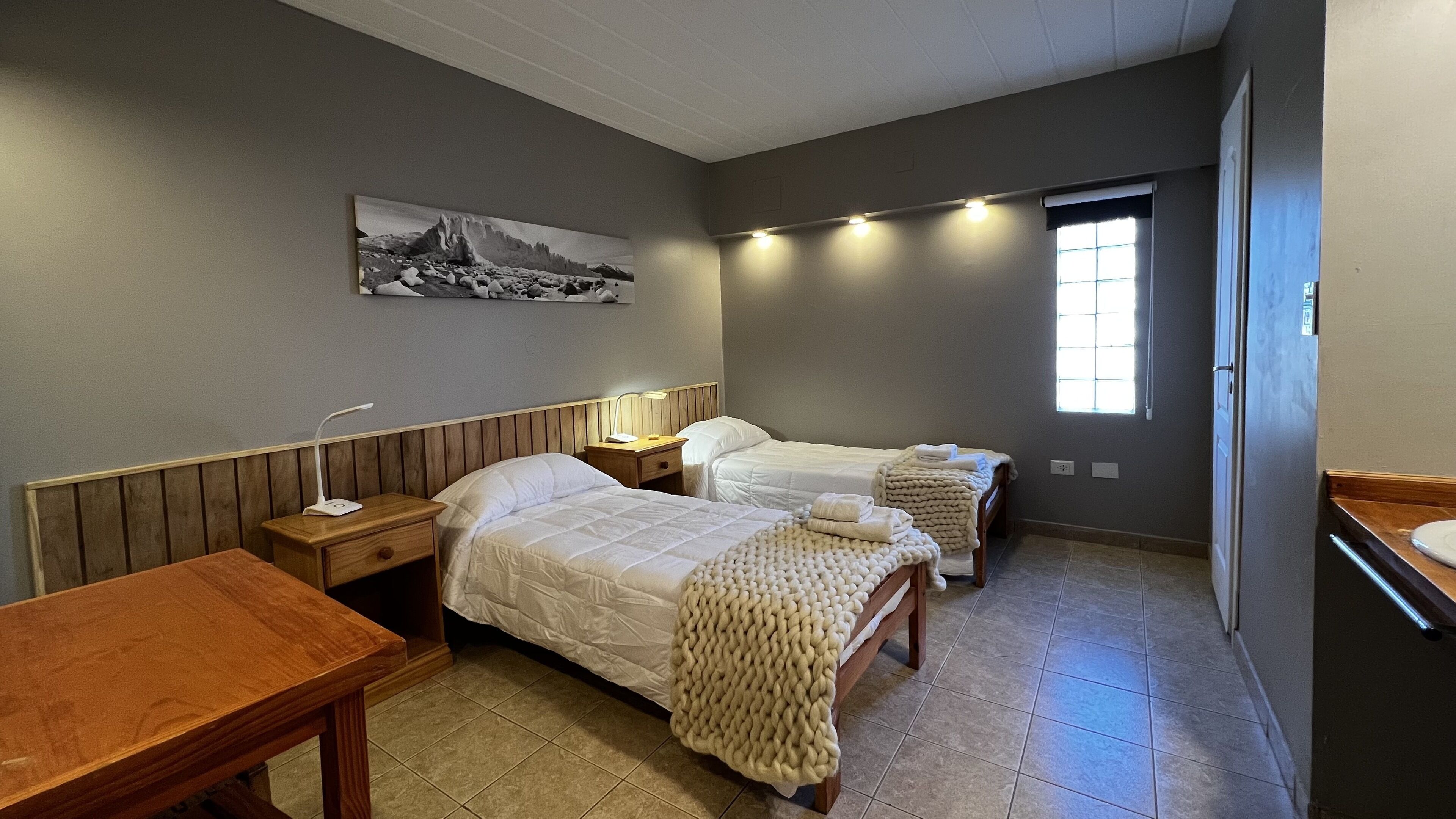 Habitación de hotel con dos camas, una mesita de noche de madera, una mesa, una lámpara de pared y una ventana con persianas.