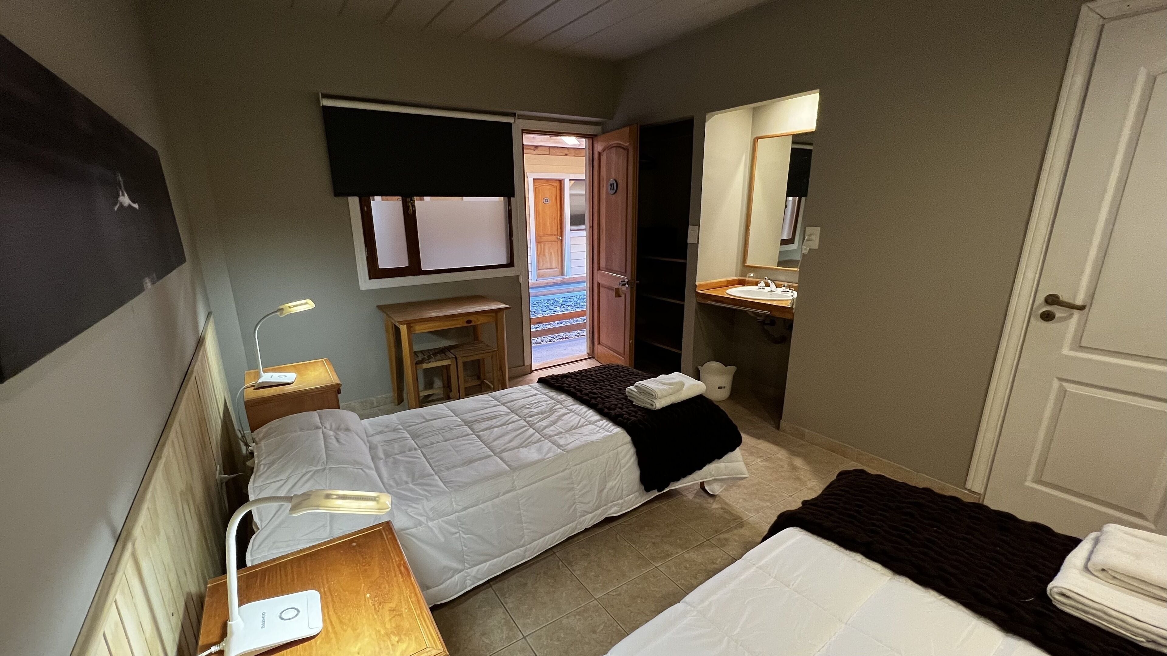 Un dormitorio pequeño y ordenado, con cama, mesita de noche, lámpara y vista al exterior a través de una puerta.